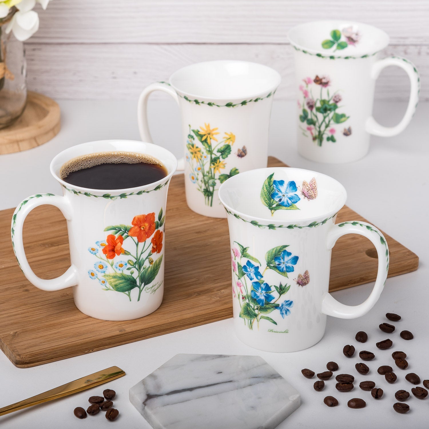 Four crest mugs on a wooden cutting board with designs of Garden Meadow flowers set on a white background with a leafy border on the mugs rim and handle. Featuring red, blue, yellow and pink flowers.
