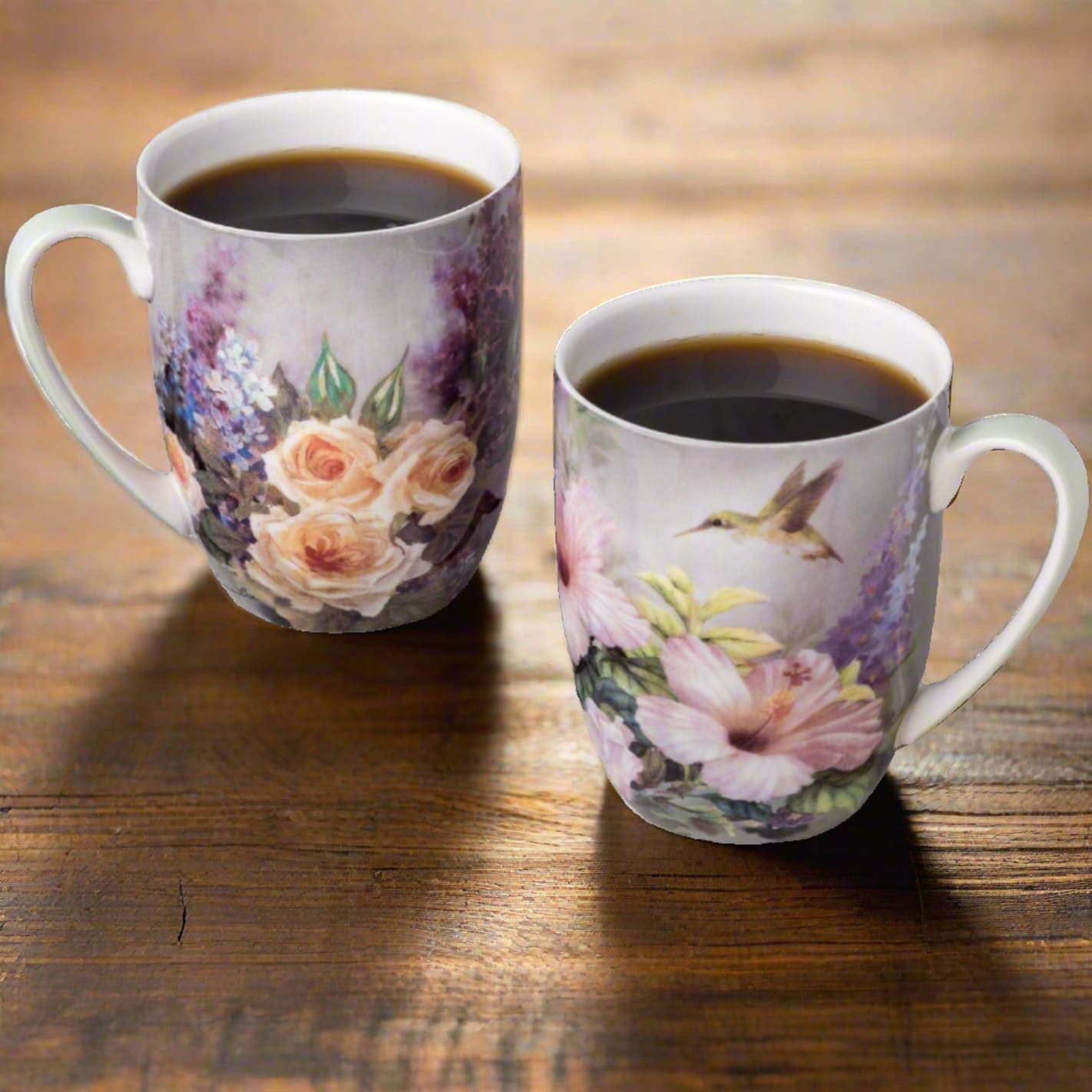 Two fine bone china mugs filled with coffee, featuring hummingbird and floral artwork with cream roses, lilacs, and pink hibiscus flowers, set on a wooden table.